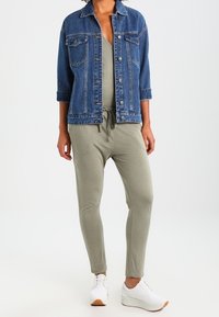 Veste en denim bleu foncé, avec un col et des poches avant, associée à un pantalon de style jogger vert olive. Les baskets sont blanches avec des semelles marron clair.