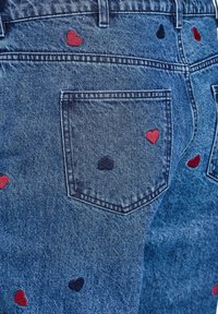 Jeans de mezclilla con una textura azul que presentan corazones bordados en rojo y azul marino en el bolsillo trasero y alrededor de las piernas.