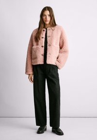Rosa Kunstpelzjacke mit einem kastigen Design, Fronttaschen und kontrastierenden schwarzen Knöpfen, kombiniert mit schwarz gesprenkelten Hosen und Schuhen.