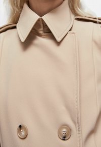 Abrigo beige de doble botonadura con un cuello estructurado, que presenta grandes botones y una textura de tela suave, con detalles de costuras visibles.