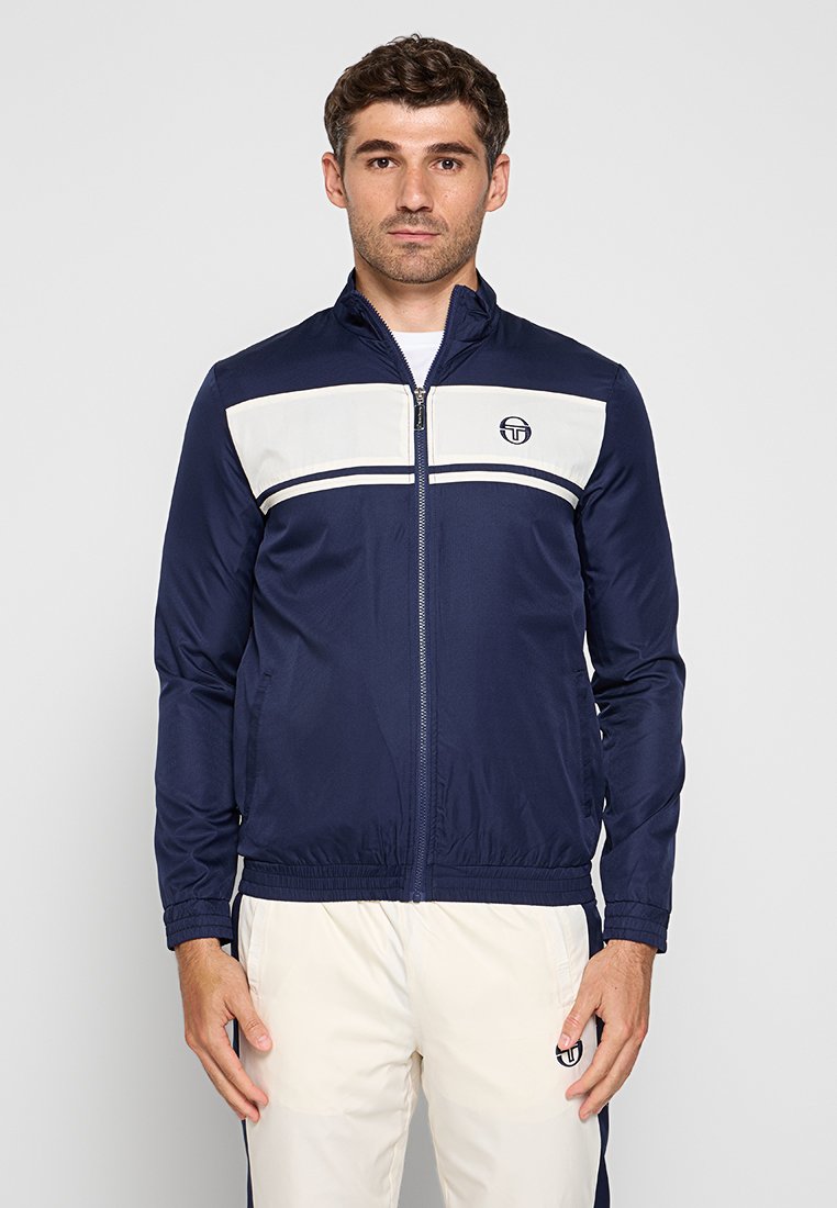 Giacca blu navy con pannello bianco e strisce sul petto; presenta una chiusura con zip e una cintura elastica, realizzata in tessuto liscio.