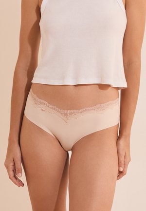 Frau trägt beige Unterwäsche mit Spitzenbesatz und ein weißes kurzes Tanktop vor einfachem Hintergrund.