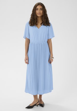 Žena s kučeravými hnedými vlasmi nosí svetlomodré plisované midi šaty s výstrihom do V a krátkymi rukávmi, v kombinácii s čiernymi slingback topánkami na nízkom podpätku.