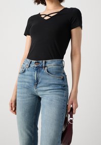 Femme portant un haut noir à manches courtes avec un col croisé et un jean taille haute bleu clair, tenant un petit sac à main violet foncé.