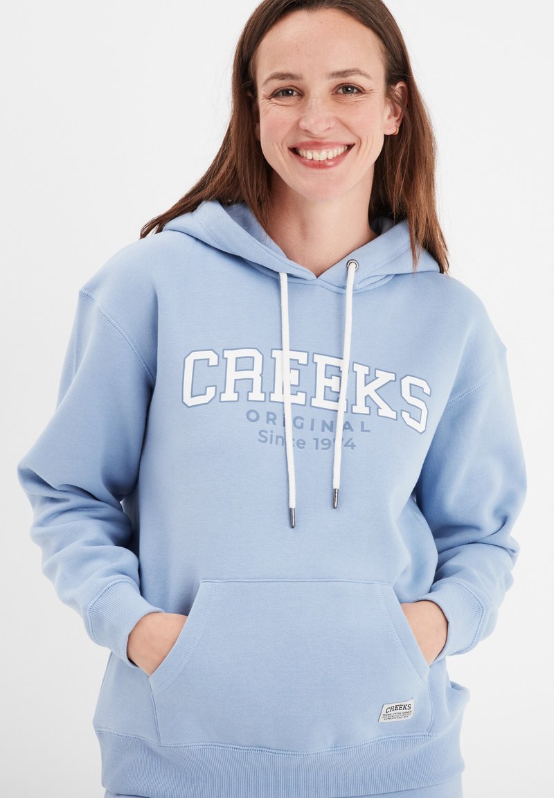 Helle blaue Kapuzenjacke mit weißem "CREEKS ORIGINAL" Text. Verfügt über eine Fronttasche, eine verstellbare Kapuze mit Kordelzug und gerippte Bündchen. Glatte Textur.