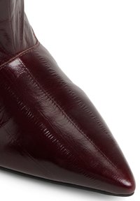 Stivale in pelle rosso scuro con punta affusolata, finitura lucida e cuciture visibili lungo la parte anteriore e laterale superiore.