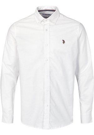 Uomo che indossa una camicia bianca con bottoni con un piccolo logo ricamato, pantaloni scuri slim, calzini bianchi e scarpe di pelle nere.