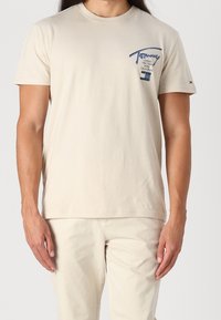 Béžové bavlněné tričko s kulatým výstřihem a krátkými rukávy, s modrým logem na levé části hrudi a jemnými švy.