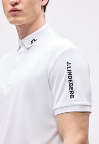 Άνδρας που φοράει λευκό πόλο μπλουζάκι με μαύρο λογότυπο JL στον γιακά και το "J.LINDEBERG" κεντημένο κάθετα στο δεξί μανίκι.