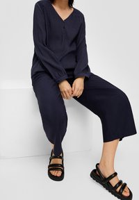 Marinblå plisserad blus med långa ärmar och V-hals, ihop med vida marinblå byxor. Svarta sandaler med remmar och tjock sula.