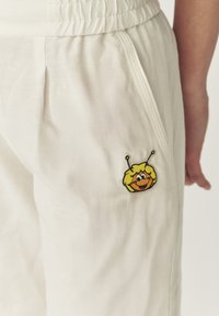 Un pantalon léger et blanc avec une taille élastique et une poche latérale, ornée d'un personnage d'abeille jaune brodé sur la jambe gauche.