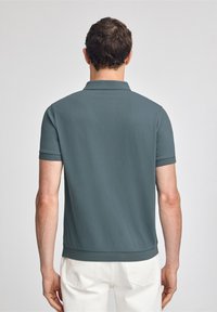Herren Poloshirt mit kurzen Ärmeln in dunklem Blaugrün, ausgestattet mit einem Kragen, gerippten Bündchen und einem geraden Saum. Glattes Material mit einer weichen Textur.