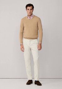 Maglione marrone chiaro in cashmere sopra una camicia a righe rosa e bianche, abbinato a pantaloni bianchi e calzature marroni.