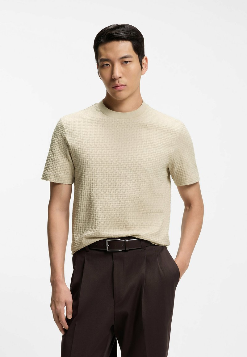 Homme portant un pull à manches courtes beige texturé et un pantalon marron foncé avec une ceinture noire, debout, une main dans la poche.
