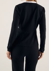 Donna che indossa un top lungo aderente nero con un colletto bianco e pantaloni neri abbinati, vista di schiena su uno sfondo semplice.