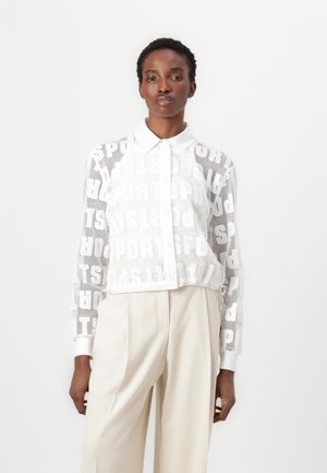White mesh jacket featuring repeating "SPORTS" text pattern, fitted design with a collar, and elastic hem. Paired with beige trousers.