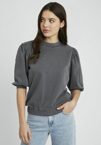 Chemise grise à manches bouffantes avec un col rond, texture côtelée et détails froncés aux manches et à l'ourlet, associée à un jean bleu clair.