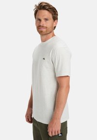 Lys grå t-shirt med en spraglet tekstur, korte ærmer og et lille logo på brystet. Modellen har olivenfarvede bukser på.