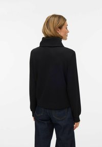 Melnā, mīkstā džempera ar augstu apkakli, vaļīgu piegriezumu un gariem piedurknēm. Kombinēts ar tumšām džinsa biksēm ar klasisku piecu kabatu dizainu.