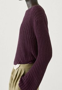 Sweter dziany w głębokim fiolecie o pudełkowym kroju. Posiada długie, szerokie rękawy i teksturowany wzór. Połączony z jasnymi spodniami.