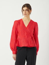 Blouse rouge à manches bouffantes avec un motif floral texturé, décolleté en V profond, et une ceinture à nouer, assortie à un pantalon sombre.