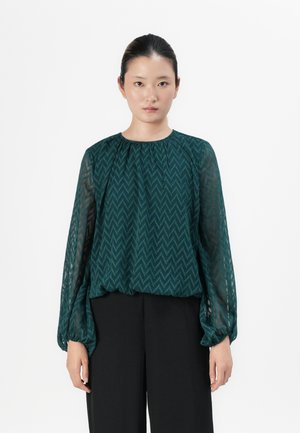 Woman stands facing forward wearing a sheer, long-sleeve green zigzag patterned blouse and black wide-leg pants against a white background.