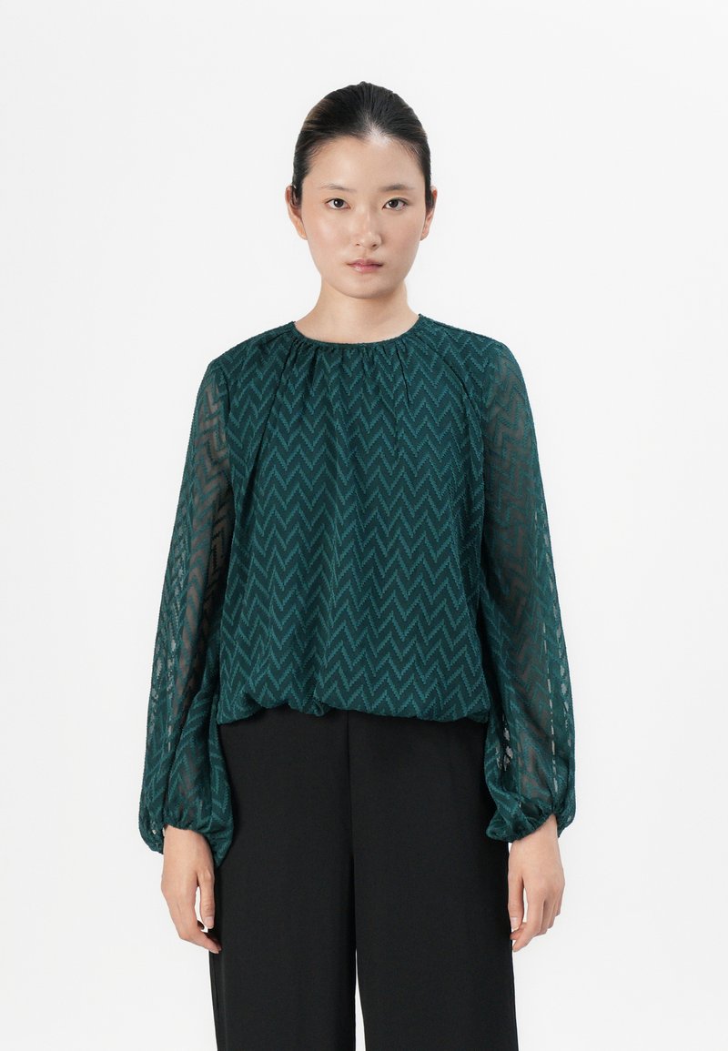 Woman stands facing forward wearing a sheer, long-sleeve green zigzag patterned blouse and black wide-leg pants against a white background.
