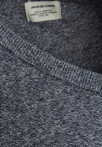 Graues Melange-Sweatshirt mit geripptem Ausschnitt, weicher Textur und einem gewebten Etikett, das den Markennamen zeigt. Nahaufnahme des Stoffdetails.