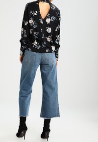 Schwarze Bluse mit Blumenmuster, langen Ärmeln und offenem Rücken, kombiniert mit hellblauen Jeans mit weitem Bein und schwarzen Stiefeln mit hohem Absatz.