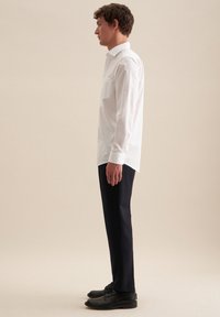 Camicia bianca a maniche lunghe con una sola tasca sul petto, abbinata a pantaloni neri su misura e scarpe nere, vista di lato.