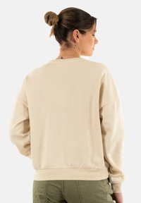 Sudadera de cuello redondo en color beige, con corte holgado, puños y dobladillo acanalados, textura suave y un diseño sencillo, mostrada desde la parte trasera.