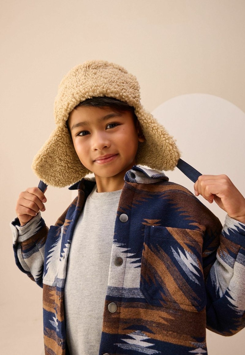 Veste en mélange de laine avec un motif géométrique bleu, marron et blanc, portant un pull gris en dessous et un chapeau beige duveteux.