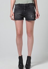 Pantalones cortos de mezclilla negra con dobladillo deshilachado, que presentan detalles desgastados y diseño de cinco bolsillos. Combinados con botas negras hasta el tobillo.