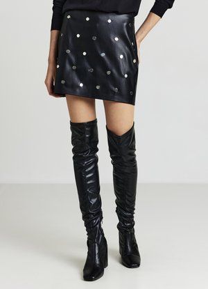 Minigonna in pelle nera con dettagli a pois metallizzati, design a vita alta, abbinata a stivali neri lucidi sopra il ginocchio. Texture liscia.