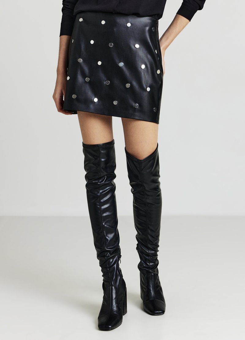 Minigonna in pelle nera con dettagli a pois metallizzati, design a vita alta, abbinata a stivali neri lucidi sopra il ginocchio. Texture liscia.