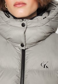 Veste matelassée grise avec capuche, dotée d'une fermeture éclair noire, de boutons-pression, et d'un petit logo "CK" noir sur la poitrine. Texture lisse et brillante.