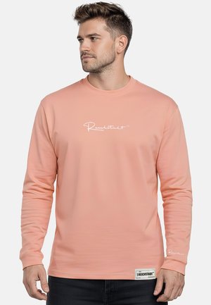 Mann mit kurzen Haaren trägt ein pfirsichfarbenes Langarmshirt mit weißem handgeschriebenem Text auf der Brust und einem Etikett nahe dem Saum, stehend vor weißem Hintergrund.