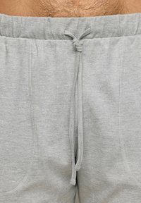 Pantaloni de trening gri deschis cu o talie elastică, dotate cu șnur de ajustare și buzunare laterale. Țesătura pare moale și texturată.