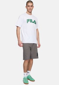 Camiseta blanca de algodón con el logo verde de FILA, combinada con pantalones cortos grises que presentan un estampado negro de FILA. El modelo lleva zapatillas verde menta y calcetines blancos.