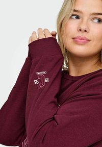Burgundijas sporta krekls ar ribotām aprocēm, uz piedurknes rakstīts "Kalifornija" un "19 95" gaiši bēšā krāsā. Mīksta auduma tekstūra, brīvs piegriezums.