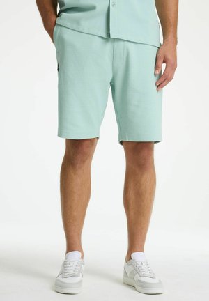 Man wearing light green textured shorts and matching short-sleeve shirt with white sneakers, standing with one hand in pocket.