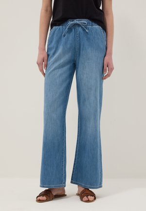 Persona con pantalones vaqueros anchos de pierna en azul claro con cintura con cordón, camiseta negra y sandalias planas marrones tejidas, de pie sobre un suelo claro.