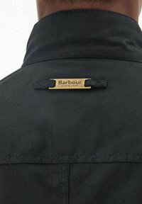 Zwarte jaskraag met een gouden metalen label gegraveerd met "Barbour since 1894" en een gestructureerde stof.