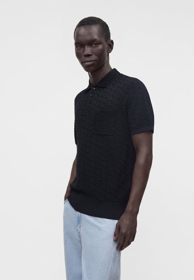 Homme portant un polo noir texturé à manches courtes avec une poche et un jean bleu clair, debout devant un fond uni.
