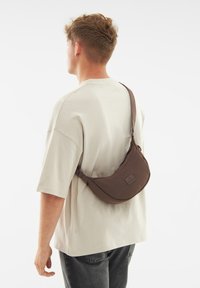 Borsa a tracolla in nylon marrone indossata sulla spalla, caratterizzata da un design curvo e da una discreta etichetta con logo, abbinata a una t-shirt oversized beige chiaro.