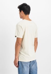 Krémové tričko s krátkým rukávem a kulatým výstřihem, vyrobené z měkké tkaniny. Na rukávu se nachází malá černá etiketa.