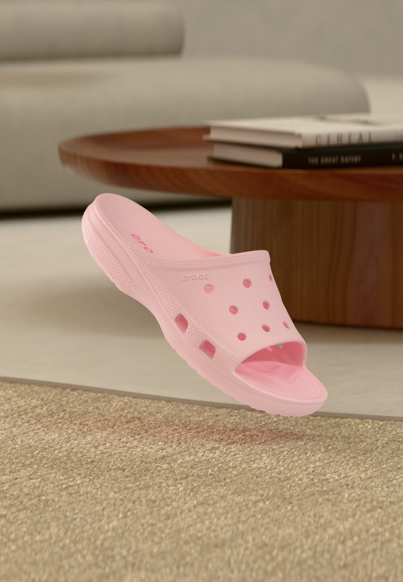 Φωτεινό ροζ clog χωρίς κορδόνια με οπές αερισμού, ακουμπισμένο σε μπεζ χαλί κοντά σε στρογγυλό ξύλινο τραπέζι με στοιβαγμένα βιβλία.