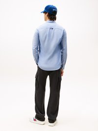 Lichtblauw overhem shirt, lange mouwen, rugzakdetail, gecombineerd met zwarte cargo pants en witte sneakers, afgemaakt met een blauwe pet.