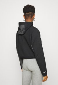 Černá krátká mikina s kapucí a síťovým vzorem, doplněná kapucí a reflexními prvky. Outfit doplňují šedé legíny a kruhové náušnice.