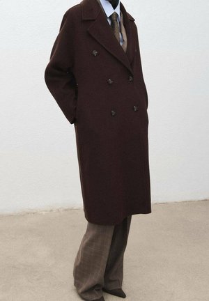 Personne portant un manteau marron foncé à double boutonnage sur une chemise bleu clair, une cravate à motifs et un pantalon marron à carreaux, debout avec les mains dans les poches.